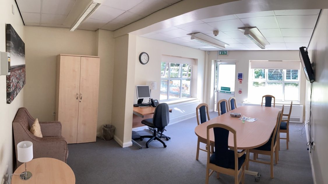 An image of a meeting room at the Lovell Park Hub. There is a wooden desk with curved edges, surrounded by chairs with wooden frames and blue upholstery. The room's carpet is grey.