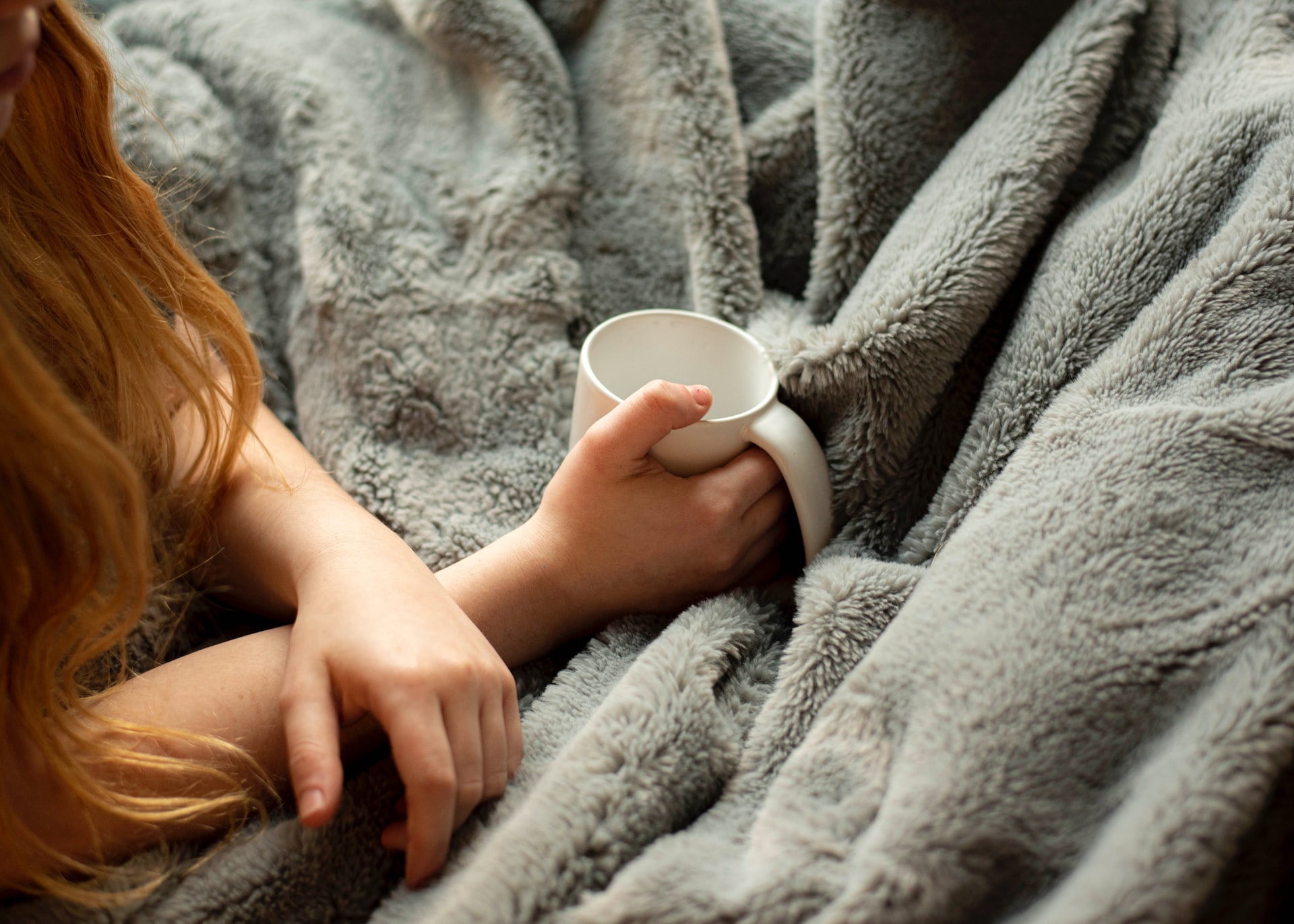 A person holding up a mug whilst being wrapped in a grey blanket