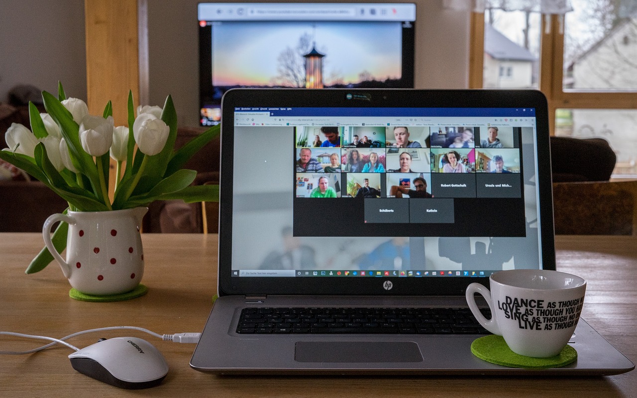 A photo of a laptop on a desk, which is showing an online meeting. There is a green coaster on top of the laptop, with a white coffee cup on top of it. To the left of the laptop, there is a USB mouse plugged in. There is also a plant pot with some white tulips growing out of it behind the mouse. All of that is on a wooden table.