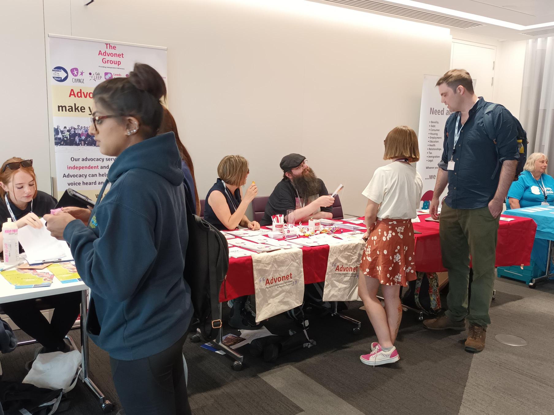 An image of members of the Leeds Autism AIM team at a stall at an information event. The stall has a red tablecloth over it, with leaflets, flyers, tote bags and other items on top of it.