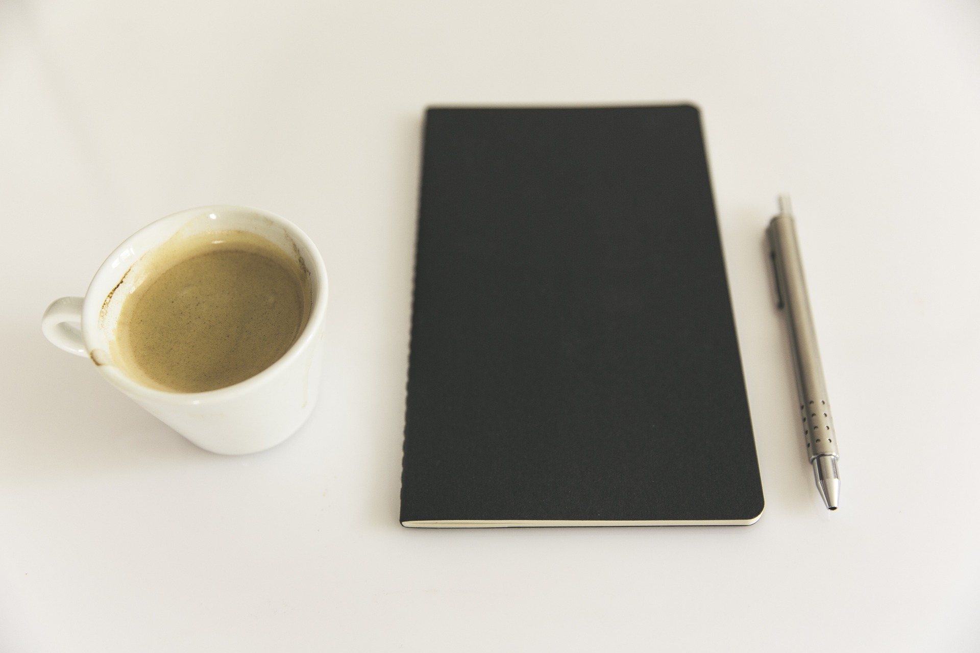 An image of a cup of coffee, a black booklet and a pen laid out on a white desk.