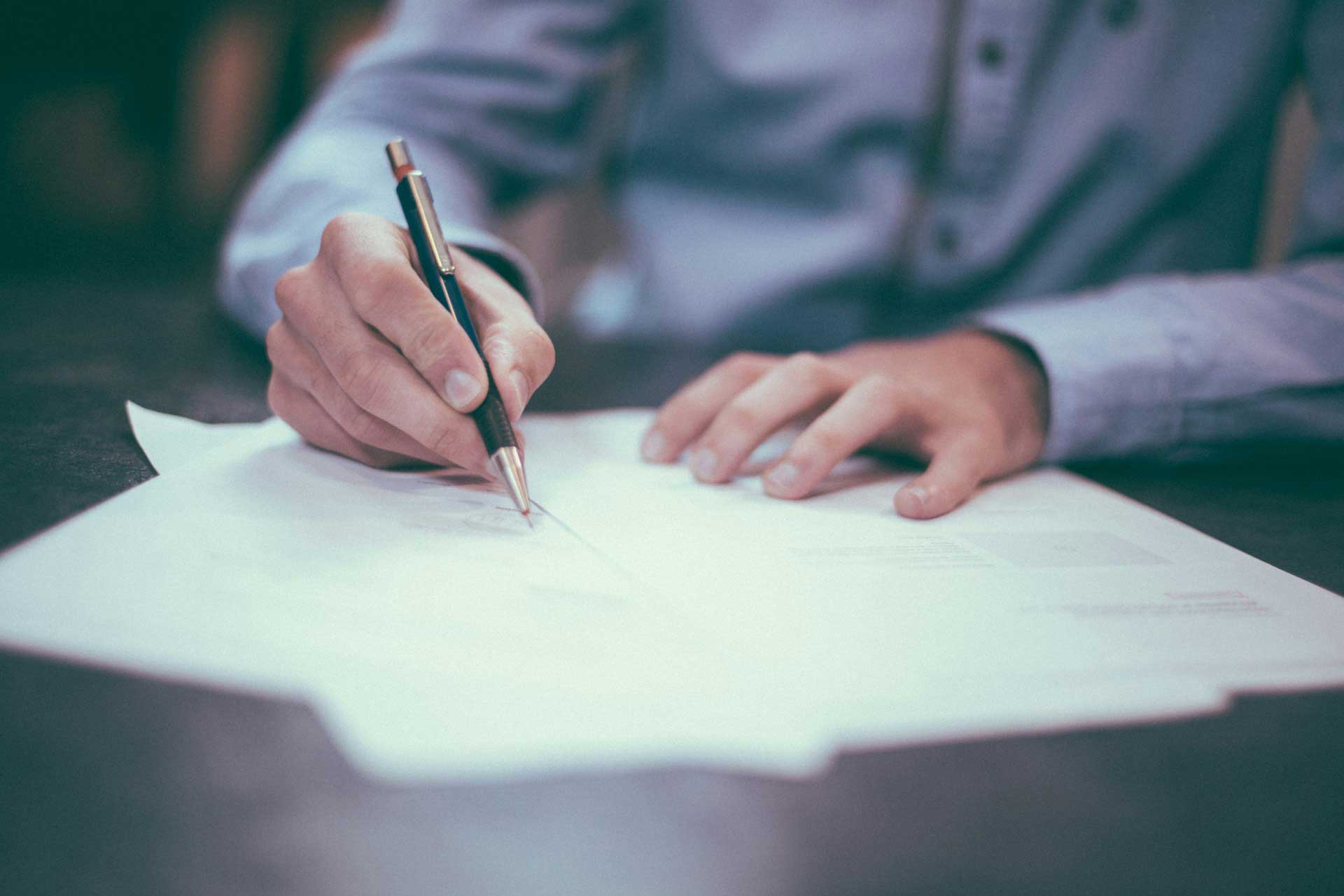 An image of someone writing on forms using a pen. They are wearing a blue-grey long-sleeved shirt and the paperwork is on a mahogany wooden table surface.