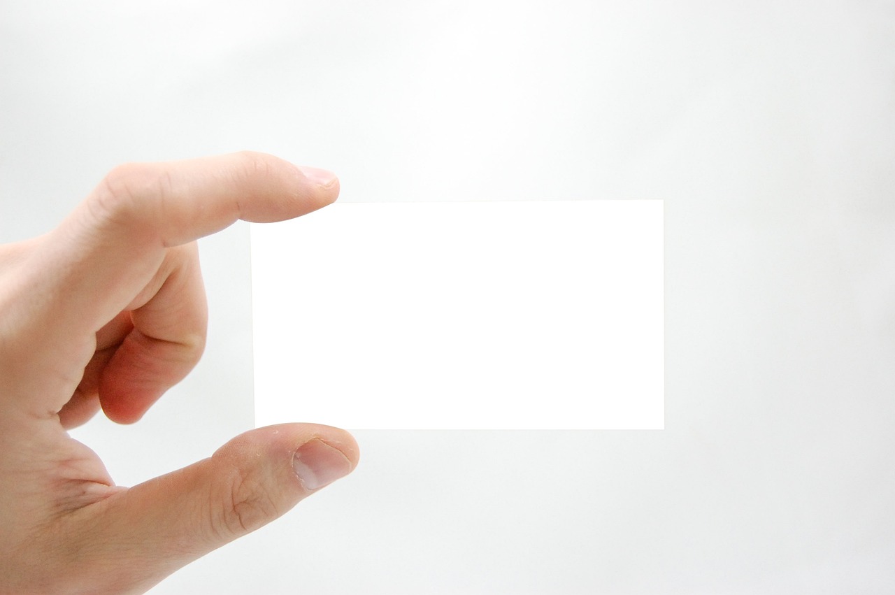 A photo of a person holding up a blank business card with their hand. The background is an off-white wall.