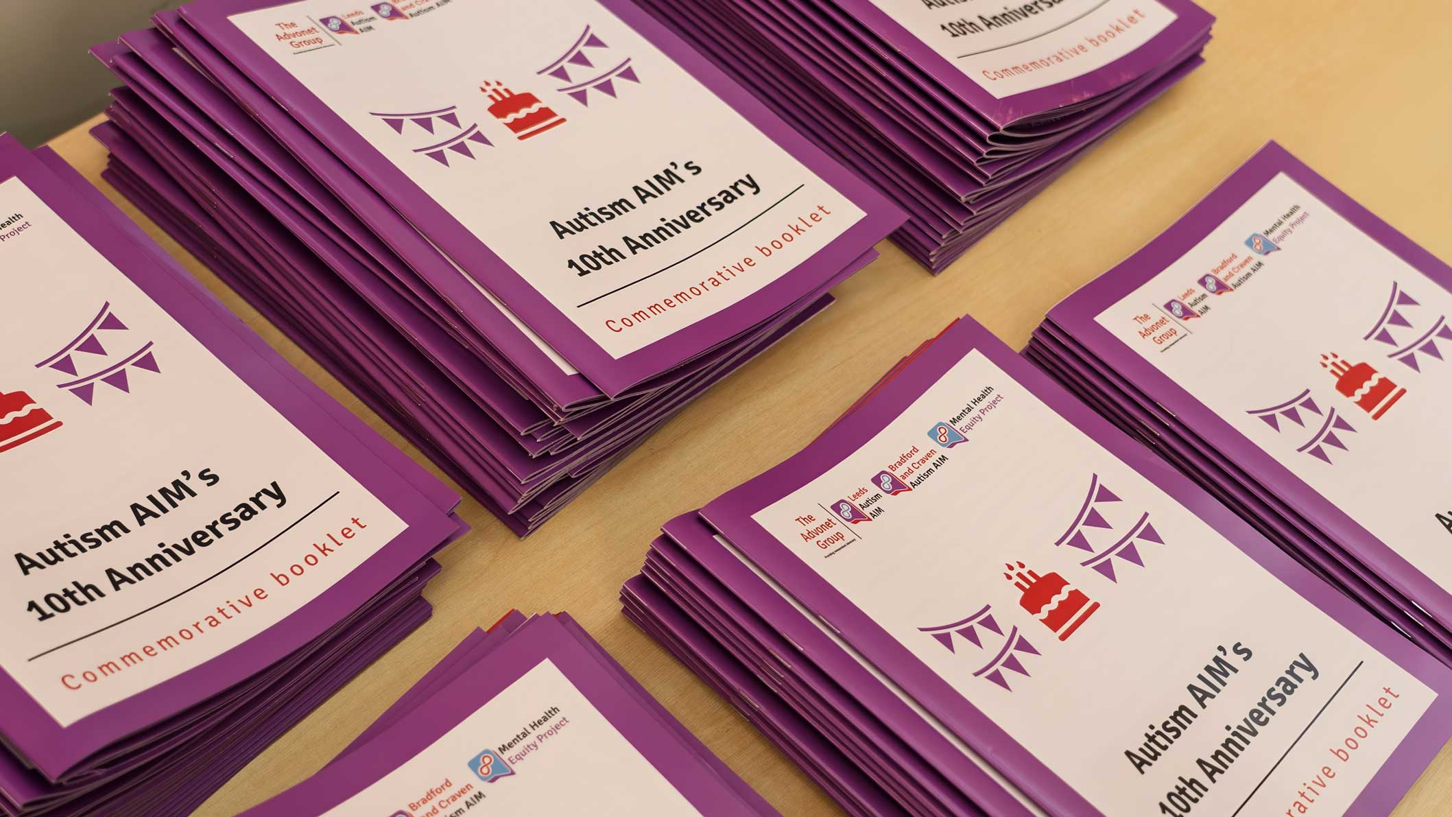 A photo of stacks of booklets marking Autism AIM's 10th anniversary, sat on a wooden table.