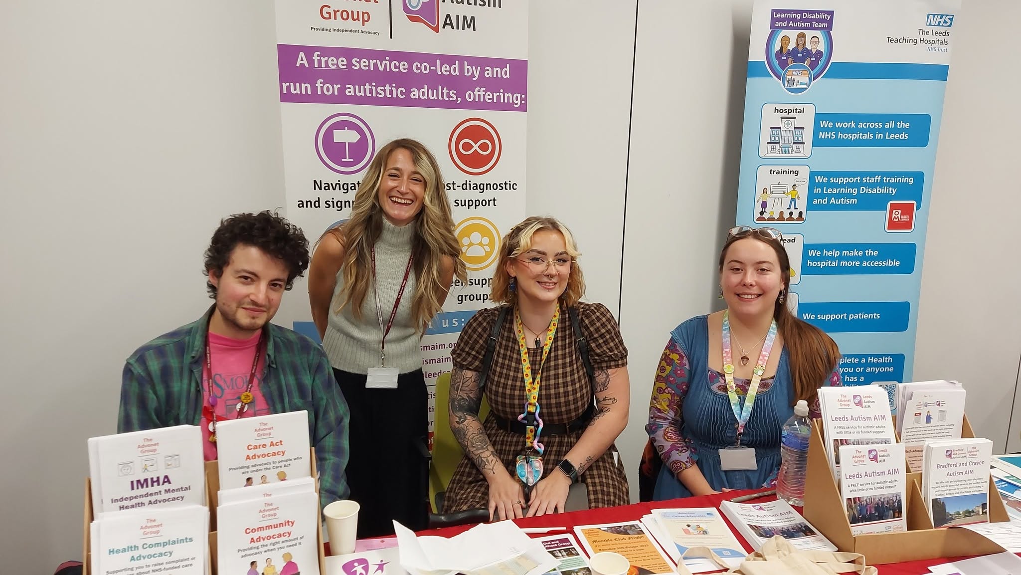 A photo of Lawrence, Danielle, Nelly and Georgia from our team at an information event. They are all behind a table with a red tablecloth and a series of leaflets and flyers on top of it.